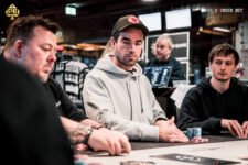 Ein Mann in einem schwarzen Anzug und einer schwarzen Sonnenbrille spielt Poker an einem Tisch in einem eleganten Casino. Er hält Karten in der Hand und schaut konzentriert auf den Tisch. Um