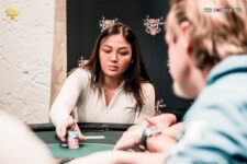 Ein Mann und eine Frau sitzen an einem Pokertisch und spielen eine Partie Poker. Der Mann hält Karten in der Hand, während die Frau konzentriert auf ihre Karten schaut. Im Hintergrund sind Pokerch