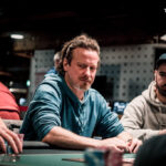 Ein Mann und eine Frau sitzen an einem Pokertisch, sie halten Karten in der Hand und schauen konzentriert auf den Tisch. Im Hintergrund sind Pokerchips und Getränke zu sehen.