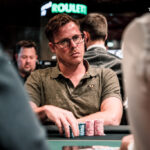 Ein Mann sitzt an einem Pokertisch und hält zwei Karten in der Hand. Er schaut konzentriert auf die Mitte des Tisches, wo verschiedene Spielchips liegen. Im Hintergrund sind weitere Spieler und
