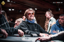 Ein Mann in einem schwarzen Anzug und einer roten Krawatte sitzt an einem Pokertisch und hält seine Karten verdeckt in der Hand, während er konzentriert auf die Karten auf dem Tisch sch