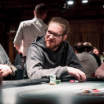 Ein Mann und eine Frau sitzen an einem Pokertisch und spielen Texas Hold'em. Der Mann hat eine Hand mit Karten und Chips vor sich liegen, während die Frau konzentriert ihre Karten betrachtet. Im Hinter