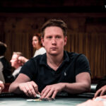 Ein Mann und eine Frau sitzen an einem Pokertisch und spielen Poker. Der Mann hat eine Hand mit Chips, während die Frau ihre Karten betrachtet. Im Hintergrund sind weitere Spieler zu sehen.