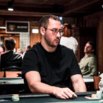 Ein Mann sitzt an einem Pokertisch und hält zwei Spielkarten in der Hand, während er konzentriert auf seine Gegner blickt. Die Tischdecke ist grün mit Pokerchips und Spielkarten