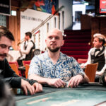 Ein professioneller Pokertisch mit grüner Filzoberfläche, umgeben von Spielern, die Karten halten und Pokerchips stapeln. Eine Mischmaschine und Getränke sind ebenfalls auf dem Tisch zu