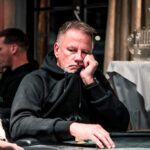 Ein Mann sitzt an einem Pokertisch und hält ein Ass und eine Herz-Dame in den Händen. Er hat eine konzentrierte Miene und schaut auf seine Karten. Im Hintergrund sind Pokerchips