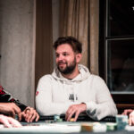 Ein grüner Pokertisch mit Spielkarten, Pokerchips und Getränken darauf, umgeben von vier Personen, die konzentriert Poker spielen.