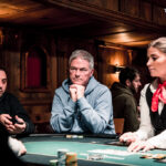 Ein professioneller Pokertisch mit grünem Filz, umgeben von Stühlen und Chips. Zwei Spieler halten ihre Karten, während der Dealer die Karten austeilt. Im Hintergrund sind weitere Spieler und eine eleg