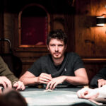 Ein Mann mit dunklen Haaren sitzt lächelnd an einem Pokertisch und hält Karten in der Hand. Er trägt ein weißes Hemd und hat eine Uhr am Handgelenk. Im Hintergrund sind