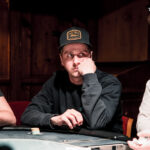 Ein Mann spielt Poker an einem Tisch mit grünem Filz. Er hält Karten in der Hand und betrachtet konzentriert seine Chips. Im Hintergrund sind weitere Spieler und eine beleuchtete Deckenle