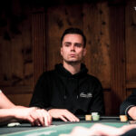 Ein Mann in einem eleganten schwarzen Anzug und einer roten Krawatte sitzt an einem Pokertisch und hält seine Spielkarten in der Hand. Um ihn herum sind Pokerchips und Gläser mit Getr