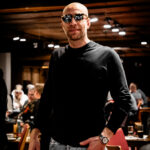 Ein Mann in einem eleganten Anzug sitzt an einem Pokertisch und hält seine Karten verdeckt in der Hand, während er konzentriert auf den Spielverlauf schaut. Um ihn herum sind Pokerchips