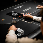 Ein Mann spielt Poker und hält seine Karten in der Hand, während er konzentriert auf den Tisch schaut. Im Hintergrund sind Pokerchips und Getränke zu sehen.