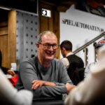 Ein Mann spielt Poker an einem eleganten Tisch in einem stilvollen Casino. Er hat Karten in der Hand und konzentriert sich auf das Spiel. Im Hintergrund sind andere Spieler und eine glamouröse Atmosphäre