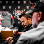 Ein Spieler mit Sonnenbrille und Hut sitzt an einem Pokertisch und betrachtet seine Karten. Um ihn herum sind Chips und Spielkarten zu sehen.