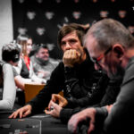 Ein Mann sitzt an einem Pokertisch und hält Karten in der Hand, während er konzentriert auf seine Gegner schaut. Die Spannung und Atmosphäre eines Pokerspiels sind spürbar.