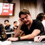 Ein Mann sitzt an einem Pokertisch und hält ein Ass und eine Pik-Dame in der Hand. Er schaut konzentriert auf seine Karten, während im Hintergrund andere Spieler und Pokerchips zu sehen sind.