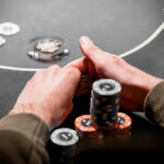 Ein Mann sitzt an einem Pokertisch und hält zwei Spielkarten in der Hand. Er schaut konzentriert auf den Tisch, umgeben von Pokerchips und einem grünen Filz. Im Hintergrund