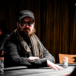 Ein Mann und eine Frau spielen Poker am Tisch, umgeben von Chips und Karten. Der Mann hat eine Hand erhoben, während die Frau konzentriert ihre Karten betrachtet.