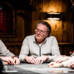 Ein Mann sitzt an einem Pokertisch mit Spielkarten und Pokerchips vor sich. Er schaut konzentriert auf seine Karten und wirkt nachdenklich. Im Hintergrund sind andere Spieler und eine Dealerin