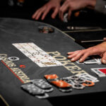 Ein Mann sitzt an einem Pokertisch in einem eleganten Casino und hält seine Karten mit einem konzentrierten Gesichtsausdruck. Andere Spieler sind im Hintergrund zu sehen, während der Croupier die