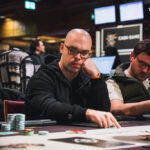 Ein Mann spielt Poker an einem eleganten Holztisch in einem stilvollen Raum mit Kronleuchterbeleuchtung. Er hält Karten in der Hand und betrachtet konzentriert den Tisch.