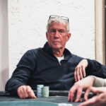 Ein Mann in einem schwarzen Anzug sitzt an einem Pokertisch und hält ein Ass und eine Pik 10-Karte in der Hand. Er hat einen ernsten Gesichtsausdruck und schaut konzentriert auf