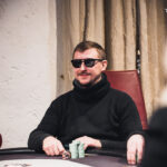 Ein Mann mit einem roten Hut und einer schwarzen Sonnenbrille spielt Poker an einem grünen Tisch mit Spielkarten und Jetons.
