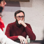 Ein Mann in einem schwarzen Anzug und einer dunkelblauen Krawatte sitzt an einem Pokertisch und hält seine Spielkarten in der Hand. Er schaut konzentriert auf den Tisch. Im Hinter