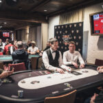 Ein Spieler hält zwei Spielkarten in der Hand und betrachtet sie konzentriert während er an einem Pokertisch sitzt. Andere Spieler und Spielchips sind im Hintergrund zu sehen.