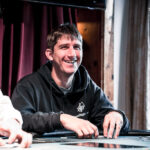 Ein Mann und eine Frau sitzen an einem Pokertisch und spielen eine Partie Poker. Der Mann hält Karten in der Hand, während die Frau nachdenklich auf ihre eigenen Karten schaut. Im Hintergrund sind Poker