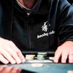 Ein Mann und eine Frau sitzen an einem Pokertisch und konzentrieren sich auf ihre Karten. Der Mann hat eine Kappe auf und die Frau trägt eine Brille. In der Mitte des Tisches liegen Pokerch