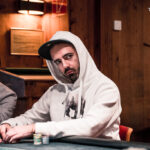 Ein Mann sitzt an einem Pokertisch und hält zwei Spielkarten in der Hand. Er schaut konzentriert auf seine Karten, während andere Spieler im Hintergrund zu sehen sind. Die Szene spielt sich in