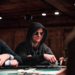 Ein Mann in einem dunkelblauen Anzug und einer weißen Hemdweste sitzt an einem Pokertisch und hält seine Karten in der Hand, während er konzentriert auf die Spielkarten auf dem Tisch