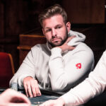 Ein Mann spielt Poker an einem Tisch mit grünen Filz und Pokerchips. Er hat eine ernste Miene und hält Karten in der Hand, während andere Spieler im Hintergrund zu sehen sind.