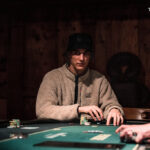 Ein Mann sitzt an einem Pokertisch und hält eine Hand mit Spielkarten. Er trägt eine dunkle Brille und konzentriert sich auf das Spiel. Andere Spieler sind im Hintergrund zu sehen.