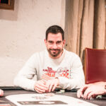 Ein professioneller Pokertisch mit grünem Filz, umgeben von Stühlen und Chips. In der Mitte des Tisches liegen Spielkarten und eine Hand hält zwei Asse. Im Hintergrund sind unschar