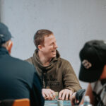 Ein Tisch mit grünem Pokertuch, auf dem Spielkarten, Pokerchips und Getränke platziert sind, umgeben von vier Personen, die konzentriert Poker spielen.