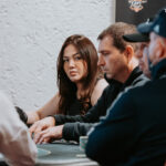 Ein Mann und eine Frau sitzen an einem Pokertisch und spielen ein spannendes Spiel. Der Mann hat eine Hand mit Karten in der Luft, während die Frau konzentriert auf ihre Karten schaut. Im Hinter