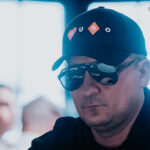 Ein Mann sitzt an einem Pokertisch und hält zwei Spielkarten in der Hand. Er hat eine konzentrierte Miene und trägt eine schwarze Kappe. Im Hintergrund sind andere Spieler und Pokerchips zu