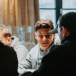 Ein professioneller Pokertisch mit grünem Filz, umgeben von Stühlen und Chips. Zwei Spieler mit Karten in der Hand sitzen am Tisch, während eine Gruppe von Zuschauern auf der anderen Seite