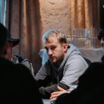 Ein Mann sitzt an einem Pokertisch und betrachtet konzentriert seine Karten, während im Hintergrund andere Spieler ebenfalls Karten halten. Die Szene strahlt eine spannende und fokussierte Atmosph