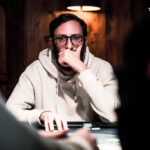 Ein Mann und eine Frau spielen Poker an einem eleganten Holztisch in einem stilvollen Casino. Der Mann hat eine Hand mit Karten und Chips vor sich, während die Frau konzentriert ihre Karten betrachtet. Die Atmos