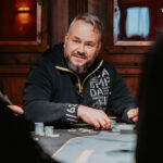 Ein Mann sitzt an einem Pokertisch, umgeben von Spielkarten und Pokerchips. Er hält seine Hand mit einem Pokerblatt in der Luft und schaut konzentriert auf den Tisch. Im Hintergrund