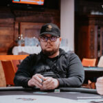 Ein Mann in einem Anzug und mit einer Krawatte sitzt an einem Pokertisch und hält zwei Spielkarten in der Hand. Er schaut konzentriert auf die Karten und wirkt ernst. Im Hinter