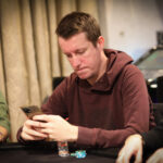 Ein Mann sitzt an einem Pokertisch, hält Karten in der Hand und schaut konzentriert auf den Tisch. In seiner Nähe sind Pokerchips gestapelt und eine Glühlampe leuchtet über