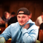 Ein Mann sitzt an einem Pokertisch in einem eleganten Casino, umgeben von anderen Spielern. Er hält Karten in der Hand und konzentriert sich auf das Spiel. Die Atmosphäre ist gespannt und glamour