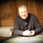 Ein professioneller Pokertisch mit grünem Filz, umgeben von Stühlen und beleuchtet von einer eleganten Lampe über dem Tisch. Karten und Chips sind auf dem Tisch verteilt, während Spieler in an
