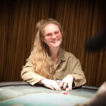 Ein professioneller Pokertisch mit grünem Filz und eleganten Holzverzierungen, umgeben von Stühlen mit roten Polstern. Spieler sitzen um den Tisch herum und halten ihre Karten während eines