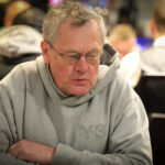 Ein Mann in einem dunkelblauen Anzug und einer weißen Hemdweste sitzt an einem Pokertisch und hält zwei Spielkarten in der Hand. Er hat einen ernsten Gesichtsausdruck und schaut